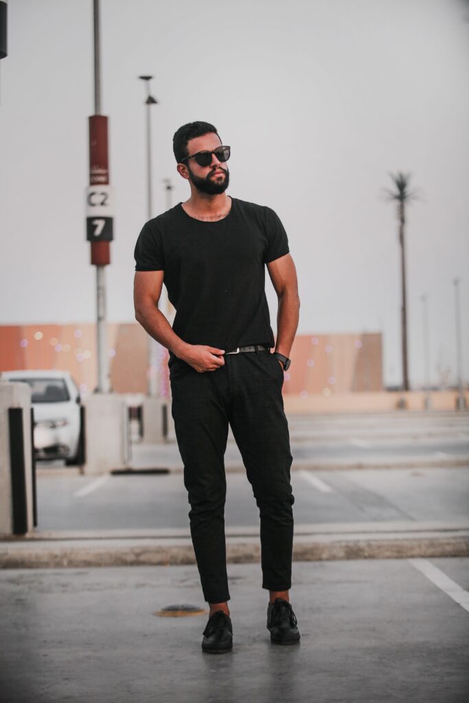 A trendy male model in black attire posing outdoors in an urban setting.